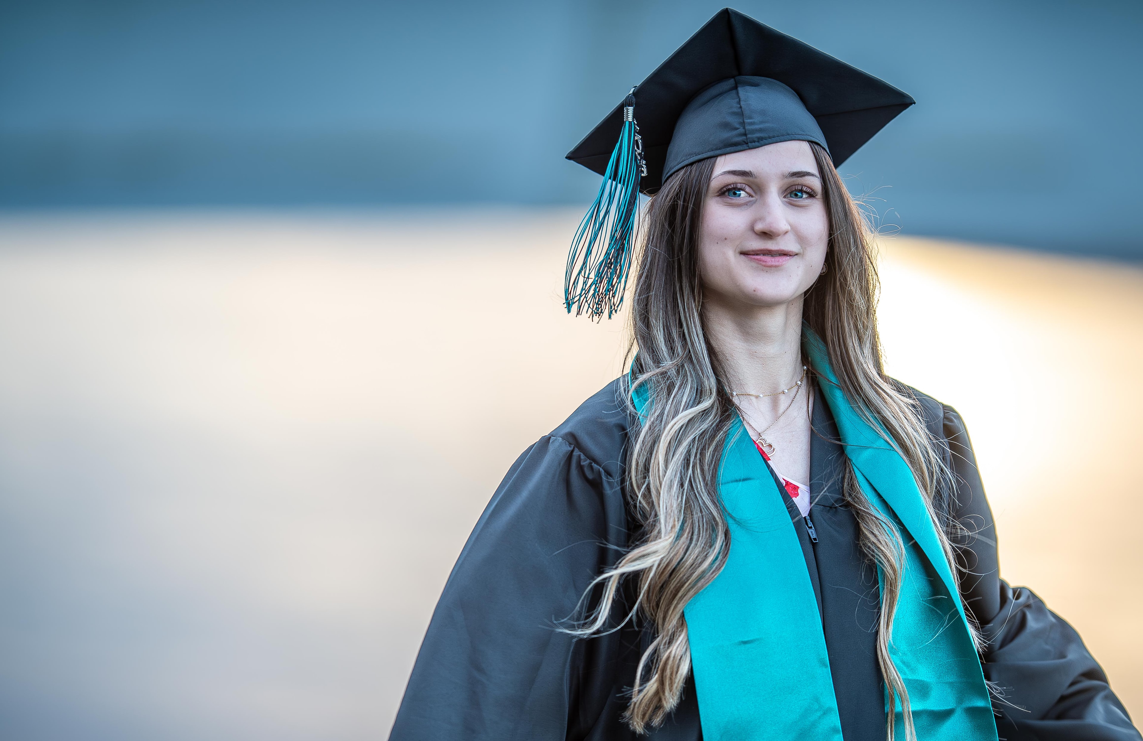 Senior portrait in graduation cap and teal stole with golden water backdrop